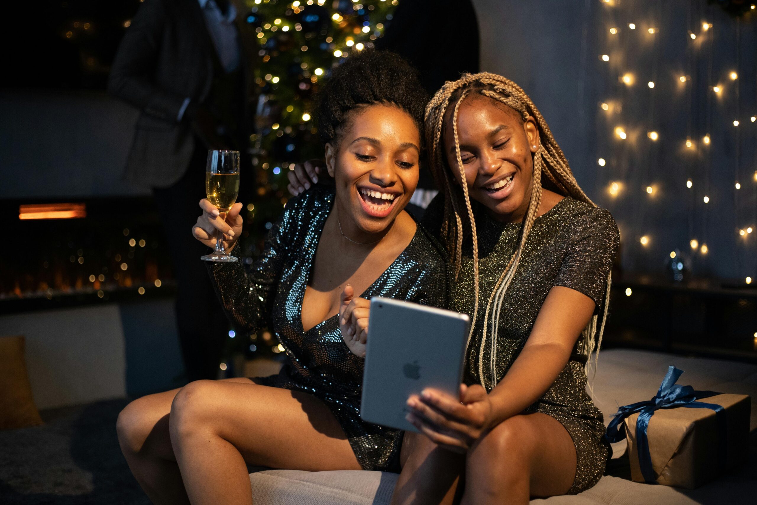 Two women enjoying a virtual holiday party with sparkly dresses and festive decorations.